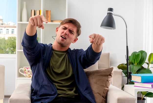 Onaangename jonge blonde knappe man zit op fauteuil wijzend op camera met twee handen in de woonkamer