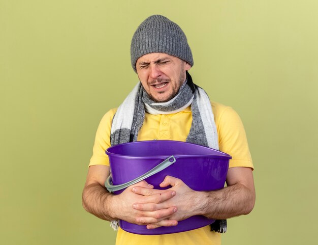 Onaangename jonge blonde zieke Slavische man met muts en sjaal houdt plastic emmer geïsoleerd op olijfgroene muur met kopie ruimte