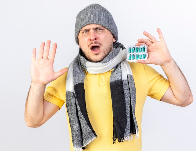 Onaangename jonge blonde zieke Slavische man met winter muts en sjaal staat met opgeheven hand en houdt pakje medische pillen geïsoleerd op een witte muur met kopie ruimte