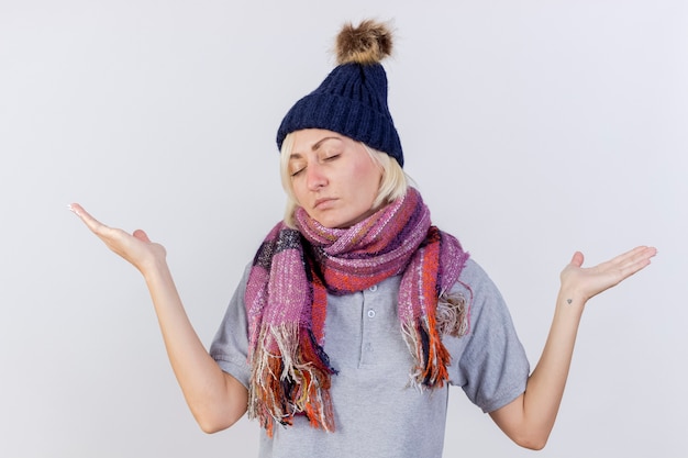 Onaangename jonge blonde zieke Slavische vrouw met muts en sjaal staat hand in hand open geïsoleerd op een witte muur met kopie ruimte