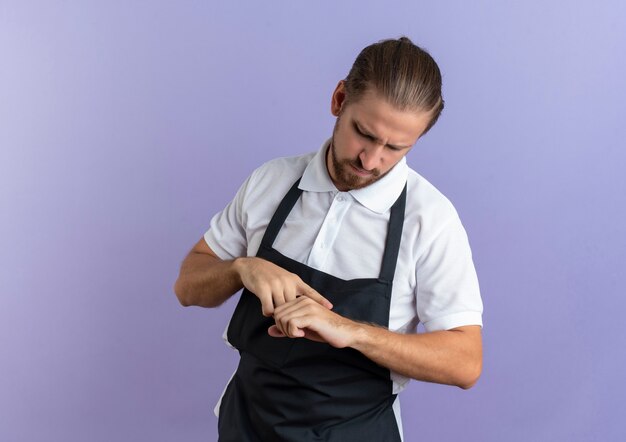 Onaangename jonge knappe kapper die uniform draagt wat zijn hand met vinger aanraakt, doet alsof kijkend naar staal geïsoleerd op paarse achtergrond met kopie ruimte