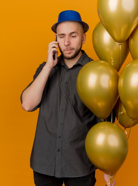 Onaangename jonge knappe Slavische feestmens met feestmuts met ballonnen praten aan de telefoon neerkijkt geïsoleerd op een oranje achtergrond