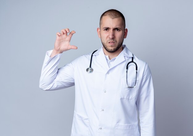 Onaangename jonge mannelijke arts die medische mantel en stethoscoop om zijn hals draagt die grootte toont die op witte achtergrond wordt geïsoleerd