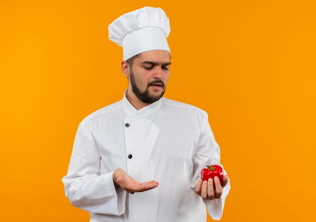 Onaangename jonge mannelijke kok in chef-kok uniforme bedrijf kijken en wijzend op peper met hand geïsoleerd op oranje ruimte