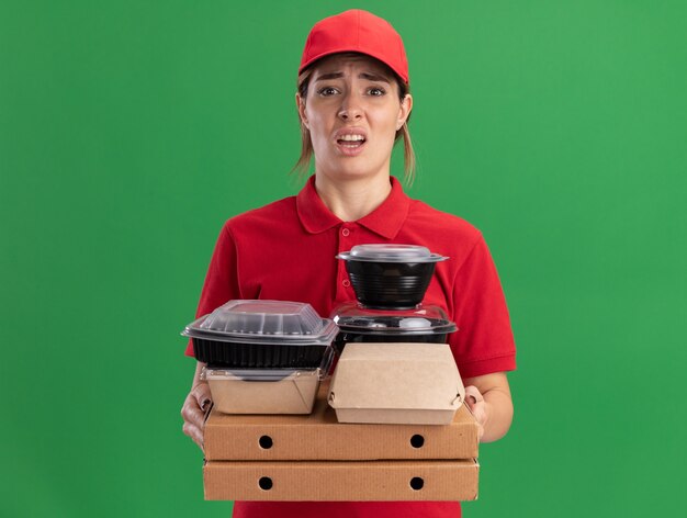 Onaangename jonge mooie bezorger in uniform houdt papieren voedselpakketten en containers op pizzadozen die op groene muur worden geïsoleerd