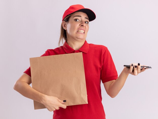 Onaangename jonge mooie bezorgvrouw in uniform houdt papieren pakket en telefoon geïsoleerd op een witte muur