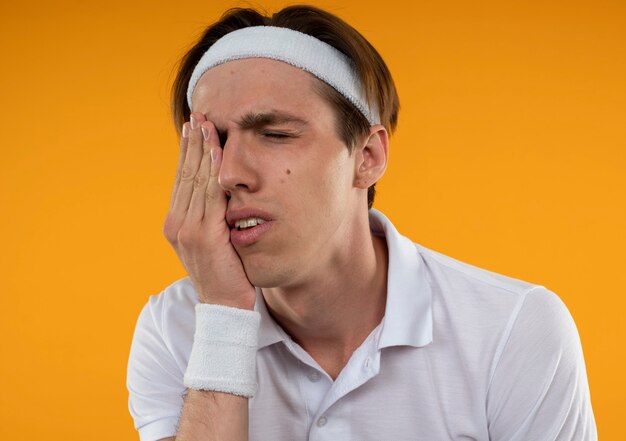 Onaangename jonge sportieve kerel met gesloten ogen die hoofdband en polsbandje dragen die hand op oog zetten dat op oranje muur wordt geïsoleerd