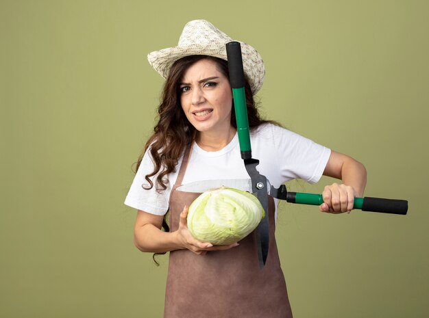 Onaangename jonge vrouwelijke tuinman in uniform draagt ?? tuinieren hoed houdt kool en tondeuse geïsoleerd op olijfgroene muur