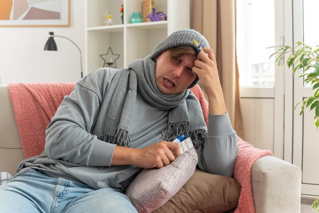 onaangename jonge zieke man met sjaal om nek met wintermuts met spuit en medicijnblisterverpakking en thermometer zittend op de bank in de woonkamer