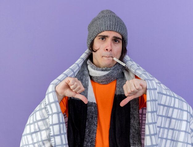 Onaangename jonge zieke man met winter hoed met sjaal gewikkeld in geruite thermometer in de mond tonen duimen omlaag geïsoleerd op paars
