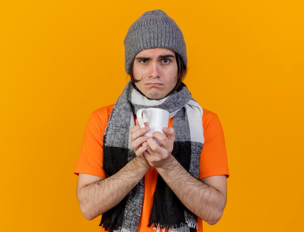 Gratis foto onaangename jonge zieke man met winter hoed met sjaal houden kopje thee geïsoleerd op een oranje achtergrond