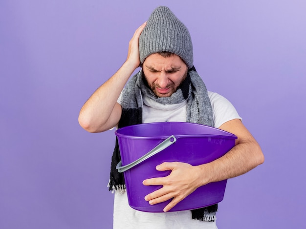 Onaangename jonge zieke man met winter hoed met sjaal met misselijkheid houden plastic busket hand zetten hoofd geïsoleerd op paarse achtergrond