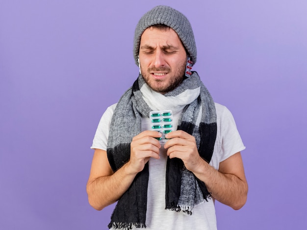 Gratis foto onaangename jonge zieke man met winter hoed met sjaal pillen onder hoed zetten en pillen geïsoleerd op paars te houden