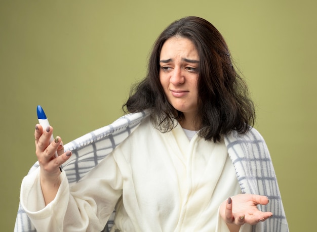 Onaangename jonge zieke vrouw die een gewaad draagt dat in geruite holding wordt verpakt en die thermometer bekijkt die lege hand toont die op olijfgroene muur wordt geïsoleerd