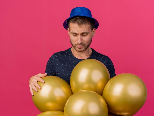 Onaangename knappe man met blauwe feestmuts houdt helium ballonnen geïsoleerd op roze muur met kopie ruimte