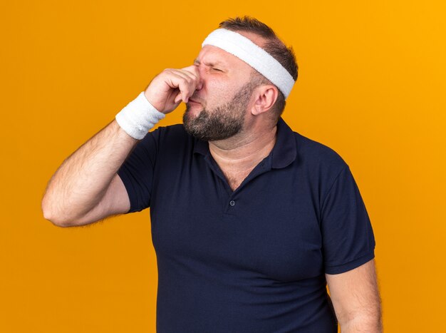 onaangename volwassen Slavische sportieve man met hoofdband en polsbandjes die zijn neus sluiten geïsoleerd op oranje muur met kopie ruimte