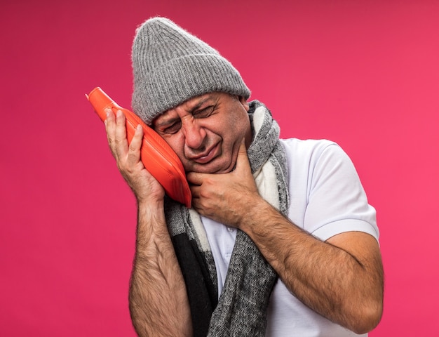 Onaangename volwassen zieke blanke man met sjaal om de nek met winterhoed legt hand op kin en houdt warmwaterkruik geïsoleerd op roze muur met kopie ruimte