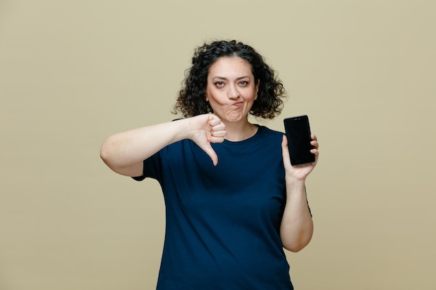 Onaangename vrouw van middelbare leeftijd die een t-shirt draagt met een mobiele telefoon die naar een camera kijkt met duim omlaag geïsoleerd op een olijfgroene achtergrond