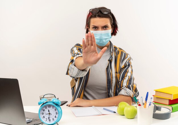 Onbehaagd jong studentenmeisje met bril op hoofd en masker zittend aan een bureau met universitaire hulpmiddelen gebaren stop bij camera geïsoleerd op een witte achtergrond