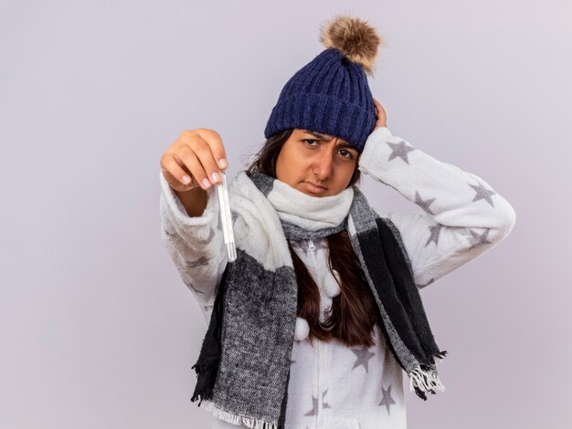 Onbehaagd jong ziek meisje dragen winter hoed met sjaal hand op het hoofd zetten en thermometer stak op camera geïsoleerd op een witte achtergrond met kopie ruimte