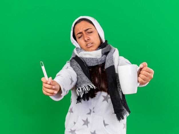 Onbehaagd jong ziek meisje met gesloten ogen kap wearin sjaal stak thermometer met kopje thee geïsoleerd op groene achtergrond te zetten