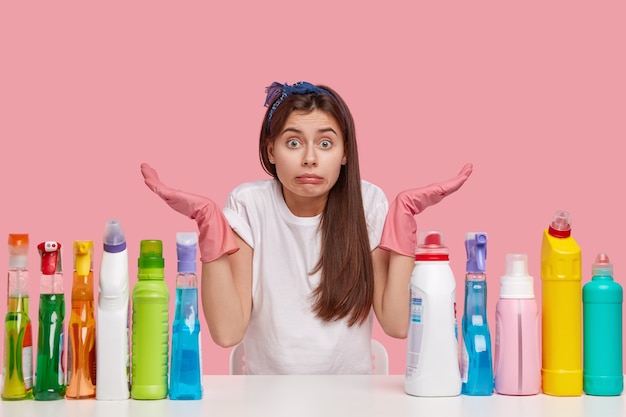 Onbewust schoonmaakmeisje draagt roze rubberen beschermende handschoenen, spreidt handen met aarzeling, poseert aan een wit bureau met schoonmaakmiddelen, kan niet beslissen welke kamer het eerst opruimt. Huishoudelijk werk concept