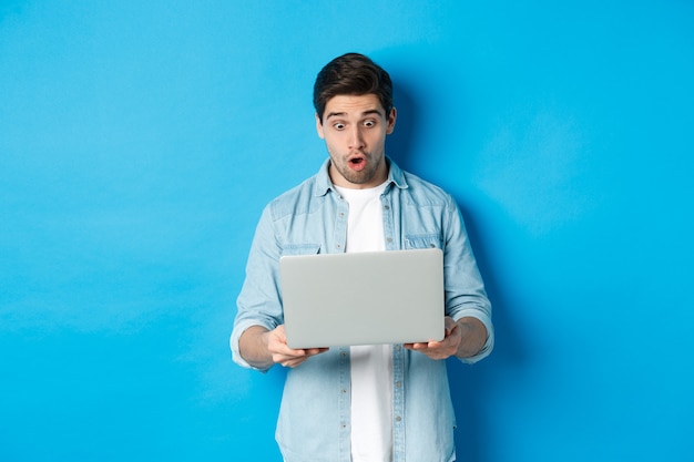 Onder de indruk blanke man die met verbazing naar het scherm van de laptop kijkt, promo op internet bekijkt, tegen een blauwe achtergrond staat