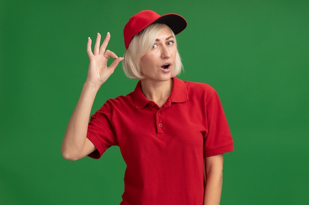 Onder de indruk blonde bezorger van middelbare leeftijd in rood uniform en pet die naar voren kijkt en ok teken doet geïsoleerd op groene muur met kopieerruimte