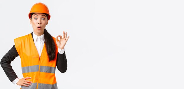 Onder de indruk en verraste Aziatische vrouwelijke bouwmanager blij met verbazingwekkende kwaliteit in veiligheidshelm en reflecterende jas met goed gebaar en zeg wow witte achtergrond
