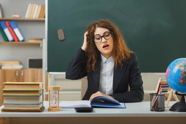 onder de indruk hand op het hoofd zetten jonge vrouwelijke leraar met een bril die een boek leest dat aan een bureau zit met schoolhulpmiddelen in de klas