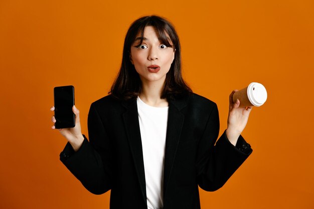 Onder de indruk houden koffiekopje met telefoon jonge mooie vrouw draagt zwarte jas geïsoleerd op oranje achtergrond