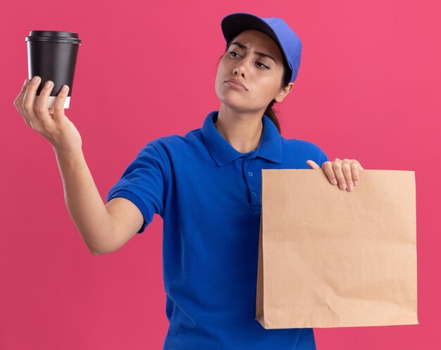 Onder de indruk jong bezorgmeisje dat uniform met pet draagt die papieren voedselpakket houdt en naar een kopje koffie kijkt in haar hand geïsoleerd op een roze muur