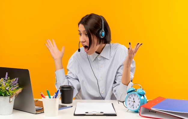 Onder de indruk jong call centreermeisje die hoofdtelefoonszitting bij bureau dragen die handen opheft en laptop bekijkt die op oranje wordt geïsoleerd