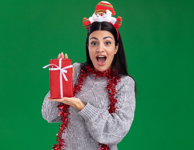 Onder de indruk jong Kaukasisch meisje dat de hoofdband van de Kerstman en een klatergoudslinger om de hals draagt die giftpakket houdt dat op groene muur met exemplaarruimte wordt geïsoleerd