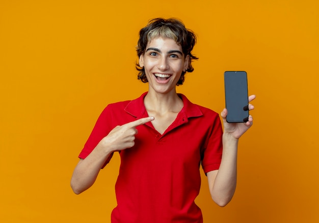 Onder de indruk jong kaukasisch meisje met pixiekapsel die en op mobiele telefoon houden richten die op oranje achtergrond met exemplaarruimte wordt geïsoleerd