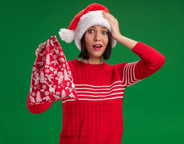 Onder de indruk jong meisje dat santahoed draagt die de zak van de Kerstmisgift houdt die hand op hoofd houdt dat op groene muur wordt geïsoleerd
