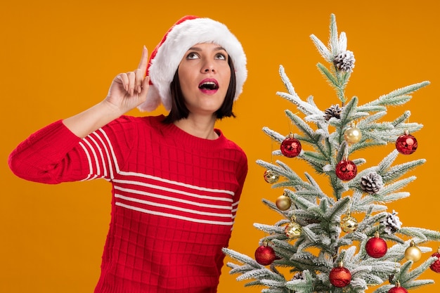 Onder de indruk jong meisje met een kerstmuts die in de buurt van een versierde kerstboom staat en naar boven wijst, geïsoleerd op een oranje achtergrond orange