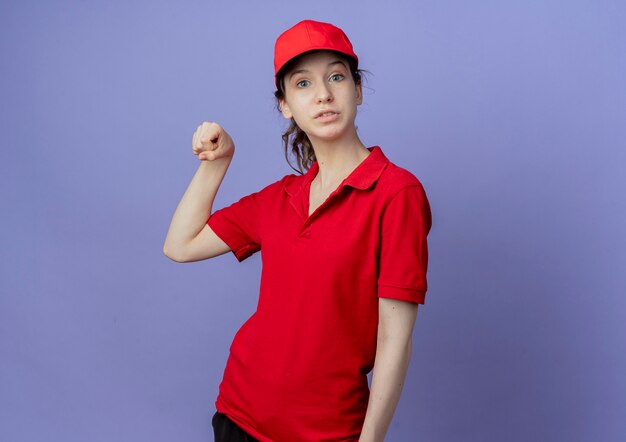 Onder de indruk jong mooi bezorgmeisje dat een rood uniform en een pet draagt, doet alsof hij iets vasthoudt dat op paarse achtergrond met kopie ruimte wordt geïsoleerd