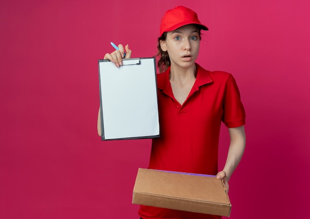 Onder de indruk jong mooi bezorgmeisje in rood uniform en pet die pizzapakketpen houdt en klembord toont dat op karmozijnrode achtergrond met exemplaarruimte wordt geïsoleerd