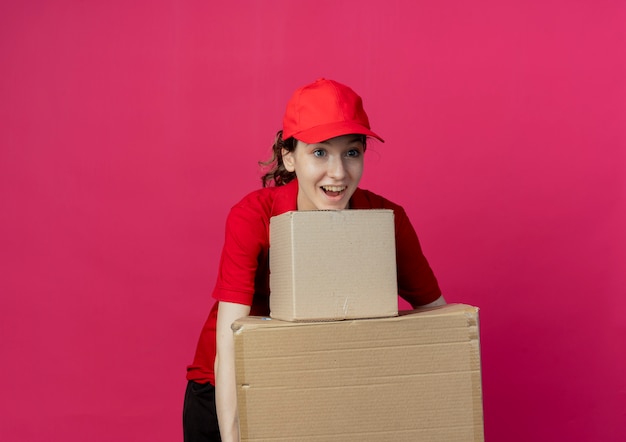 Onder de indruk jong mooi bezorgmeisje in rood uniform en pet die recht op zoek zijn met kartonnen dozen geïsoleerd op een karmozijnrode achtergrond met kopie ruimte