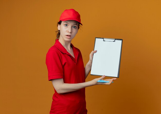 Onder de indruk jong mooi leveringsmeisje in rood uniform en GLB die pen houden en klembord tonen dat op oranje achtergrond met exemplaarruimte wordt geïsoleerd