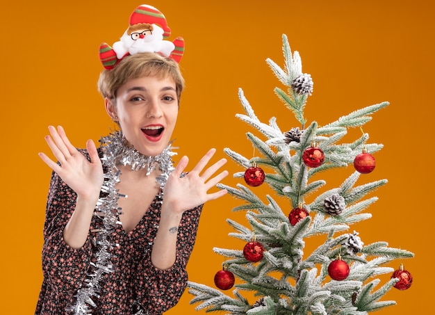 Onder de indruk jong mooi meisje draagt ?? de hoofdband van de kerstman en klatergoud slinger rond de nek staan ?? in de buurt van versierde kerstboom met lege handen geïsoleerd op een oranje achtergrond