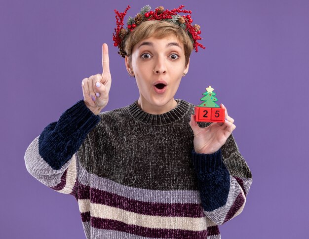Onder de indruk jong mooi meisje draagt ?? kerst hoofd krans bedrijf kerstboom speelgoed met datum omhoog geïsoleerd op paarse muur