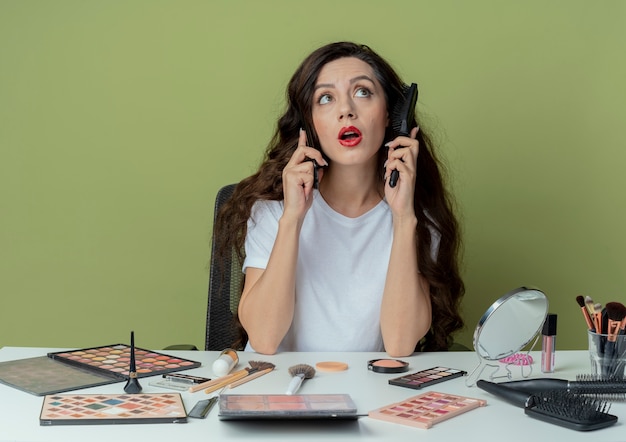 Onder de indruk jong mooi meisje zit aan make-up tafel met make-up tools hoofd aan te raken met kam kant kijken en praten over telefoon geïsoleerd op olijfgroene achtergrond