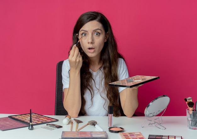 Onder de indruk jong mooi meisje zittend aan de make-up tafel met make-up tools oogschaduw palet houden en oogschaduw toe te passen met oogschaduw borstel geïsoleerd op crimson achtergrond