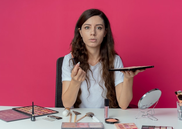 Onder de indruk jong mooi meisje zittend aan make-up tafel met make-up tools oogschaduw palet en borstel geïsoleerd op een karmozijnrode achtergrond te houden