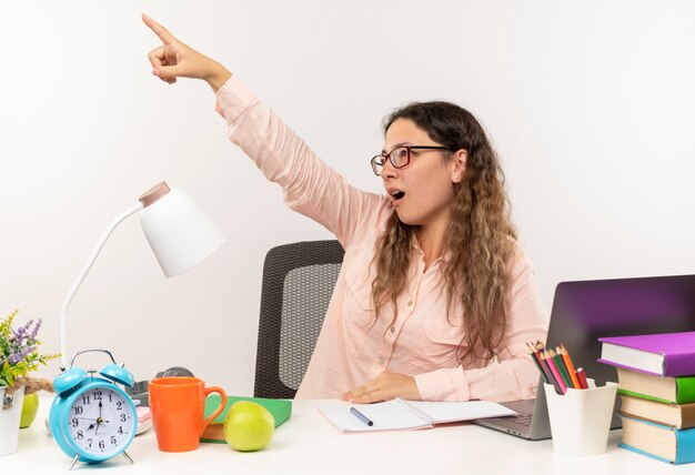 Onder de indruk jong mooi schoolmeisje die glazen dragen die aan bureau met schoolhulpmiddelen zitten die haar huiswerk doen kijken en naar kant wijzen die op wit wordt geïsoleerd