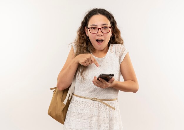 Onder de indruk jong mooi schoolmeisje die glazen en achterzak dragen die en op mobiele telefoon houden die op wit wordt geïsoleerdd