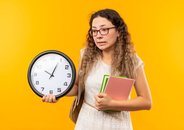 Onder de indruk jong mooi schoolmeisje die glazen en achterzak dragen die klok notitieblok en boek houden die kant bekijken die op geel wordt geïsoleerd