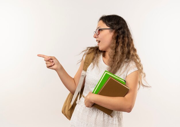 Onder de indruk jong mooi schoolmeisje die glazen en rugtas dragen die boeken loooking en naar kant richten die op wit wordt geïsoleerd